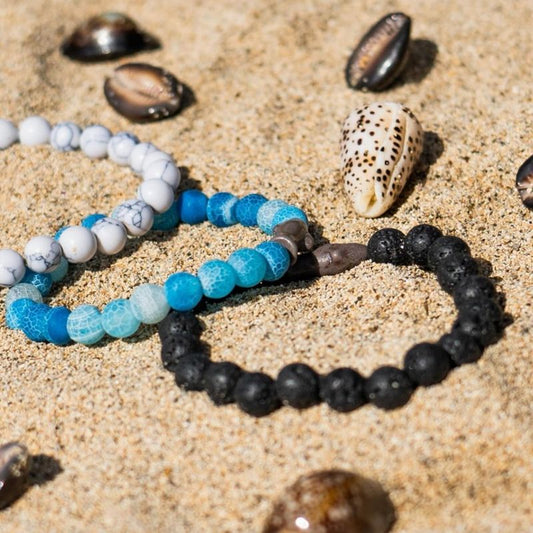 Three beaded fahlo bracelets in blue, black, and white with seal charms on sand with shells around