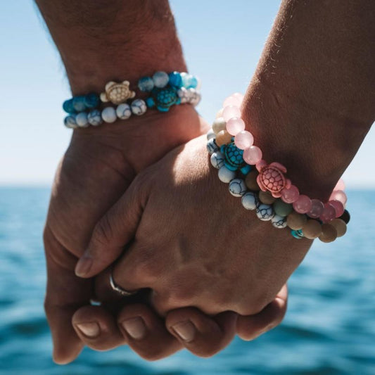 Two hands holding each other with beaded fahlo bracelets with turtle charms against a blue sky and water background