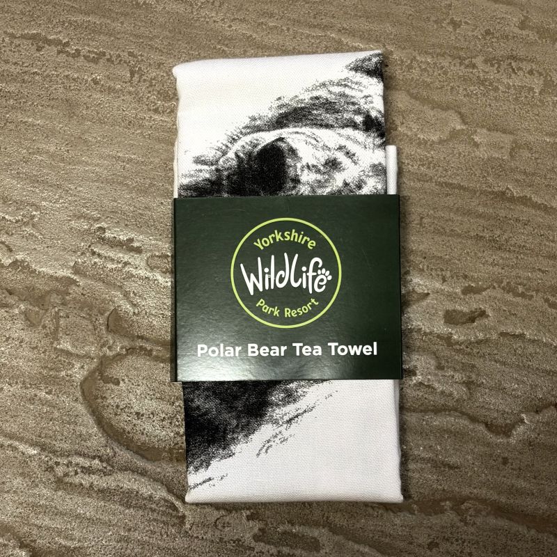 A folded Yorkshire Wildlife Park Resort tea towel with a black and white polar bear print, wrapped in a dark green paper band labeled 'Yorkshire Wildlife Park Resort Polar Bear Tea Towel', placed on a textured beige stone surface.