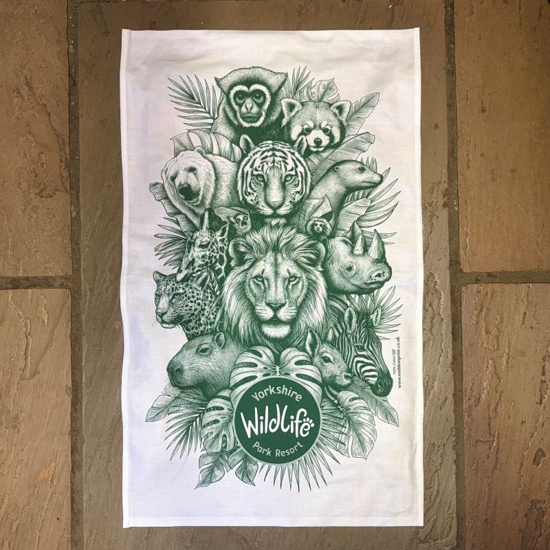 A white tea towel featuring a detailed green and white image of several animals at the yorkshire wildlife park displayed flat against a rustic brown flagstone background.