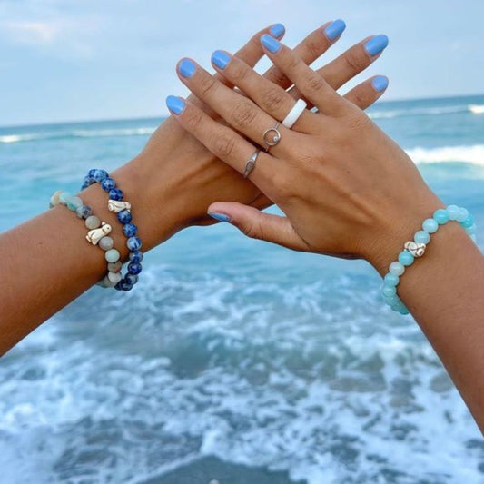 Two hands with blue nail polish and three fahlo bracelets with penguin charms against a beach background