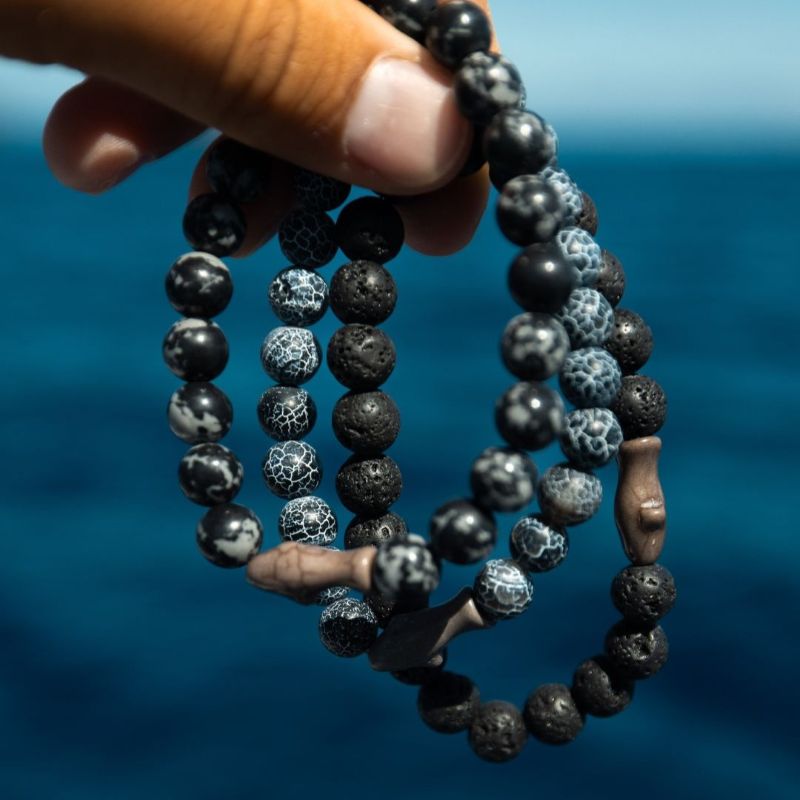 Black gray and blue beaded fahlo bracelets with shark charms held by a hand with a blue background