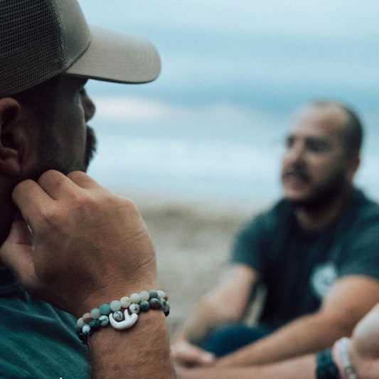 Two men sitting on a beach, one wearing a cap and fahlo bracelets with sloth charms.