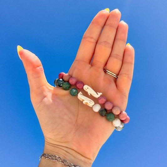 Hand holding two beaded bracelets with whale charms against a clear blue sky